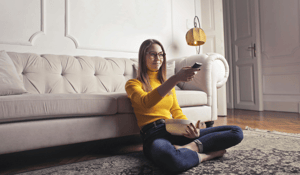 Woman sitting in yellow swaeter pointing tv remote at tv sitting up against a cream couch 